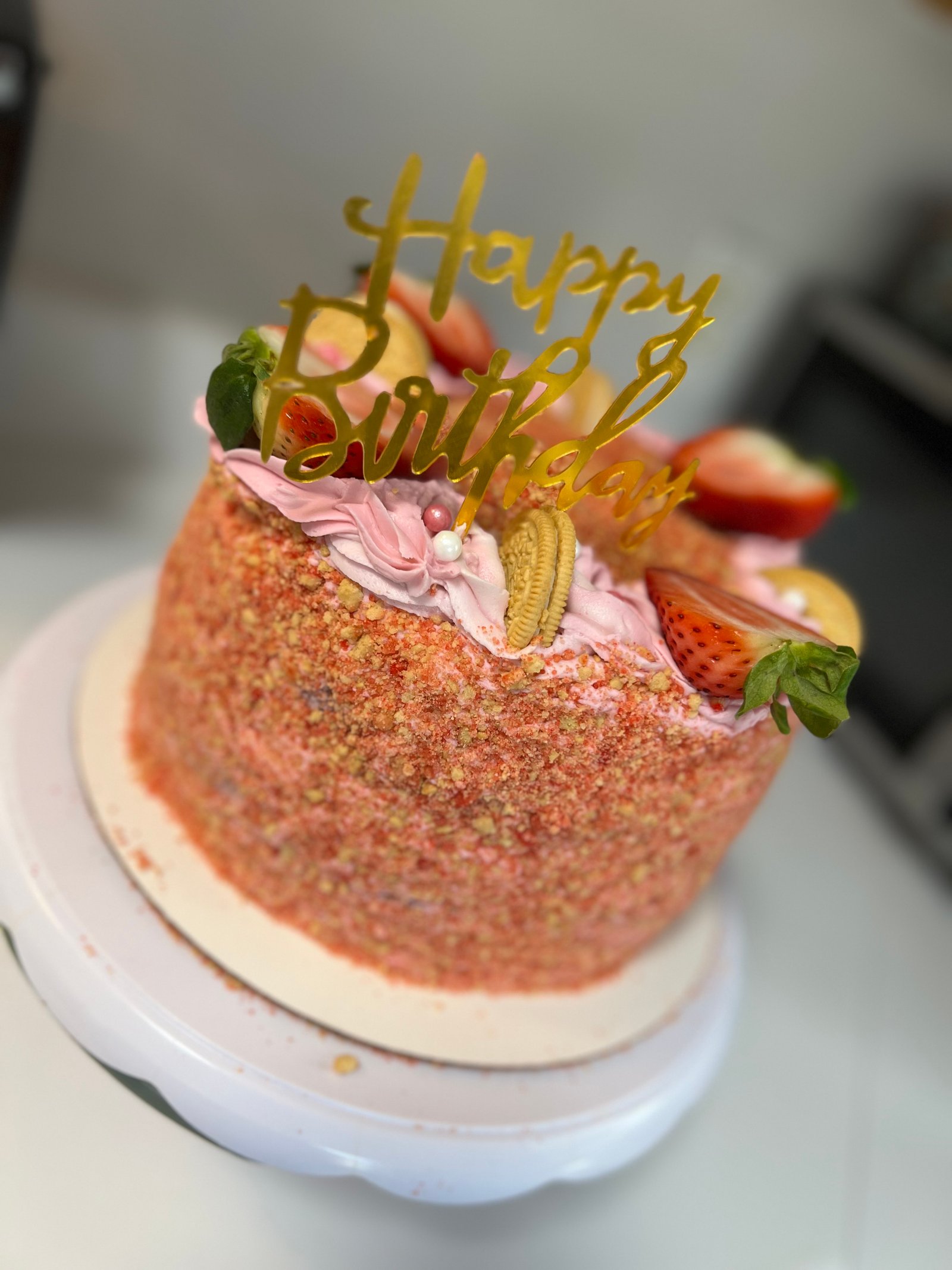Strawberry crunch birthday cake with pink frosting, fresh strawberries, and gold topper